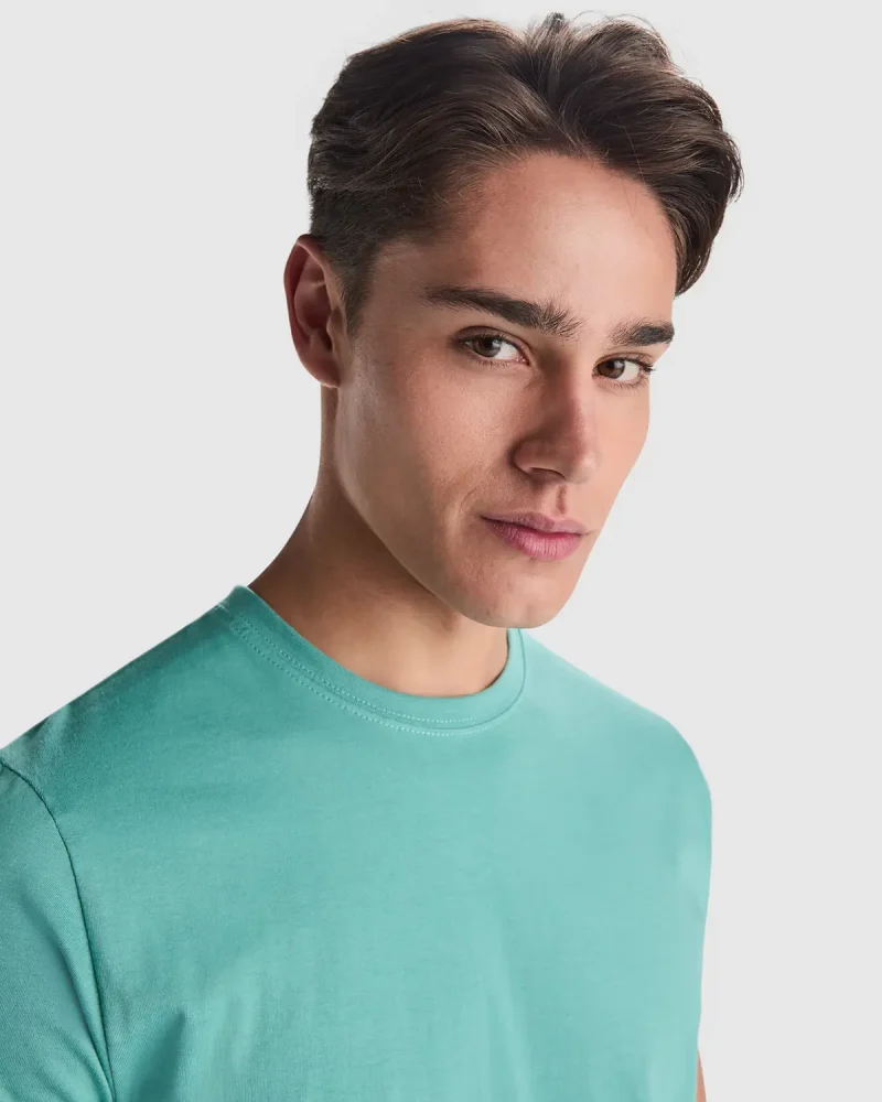 Young man with short brown hair wearing a turquoise crew-neck T-shirt, photographed from the chest up against a light background.